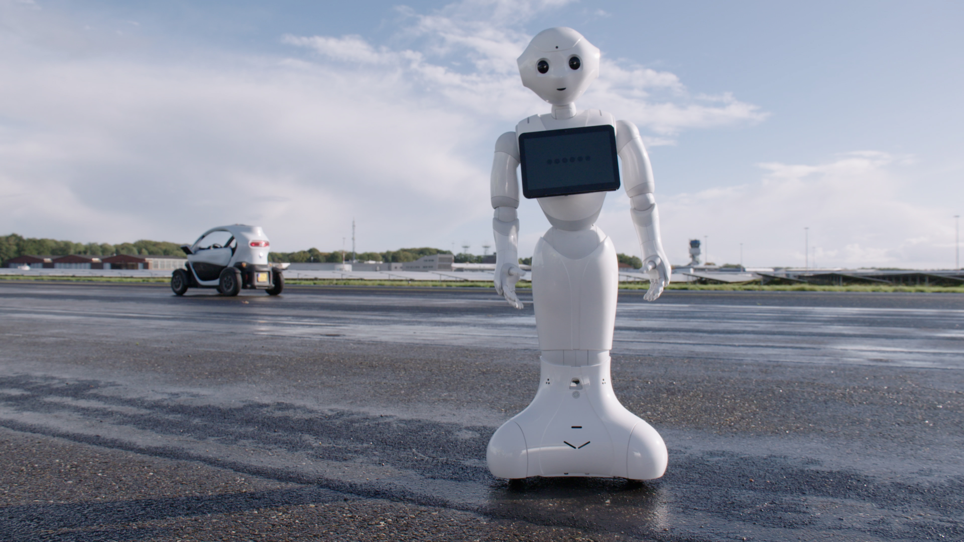 Een witte robot met een zwart tv scherm op een weg met op de achtergrond een kleine witte auto