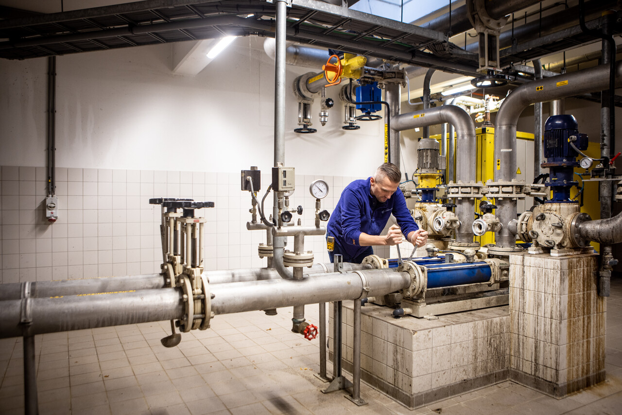 Een monteur in blauwe werkkleding werkt met gereedschap aan een installatie in een technische ruimte vol leidingen, afsluiters en meters. De ruimte is betegeld en goed verlicht, met boven hem een kabelgoot en rondom verschillende pompen en motoren. De focus ligt op onderhoud of reparatie van een buizensysteem.