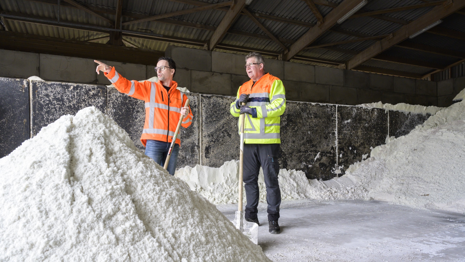Twee mensen in een oranje jas staan bij een berg met zout, waarbij de één de ander iets aanwijst.