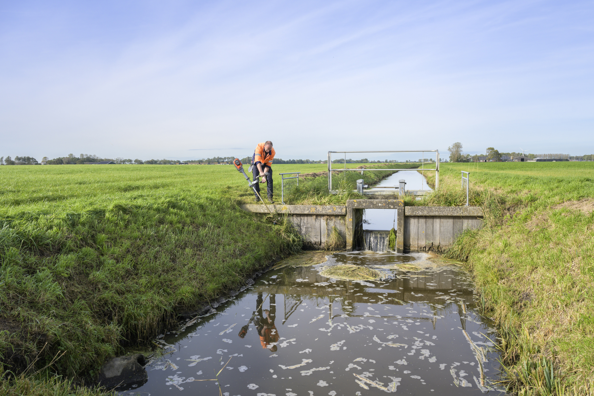 Een man is in werkkleding met een snoeimachine gras aan het snoeien bij een water doorlaat.